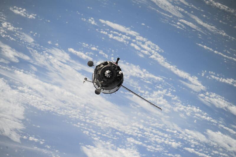 A satellite approaching the viewer, captured from an aerial perspective, with a backdrop of blue sky and clouds below