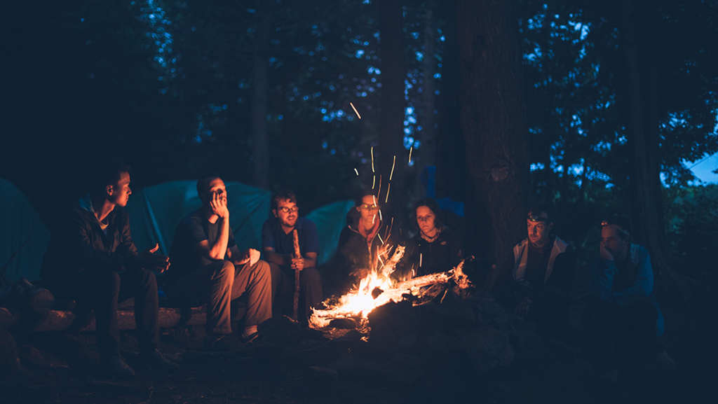 A group of people sitting by a campfire at night with tents in background
