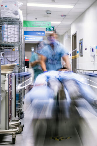 Blurry image of staff in blue scrubs wheel a patient on a stretcher through the Emergency Department