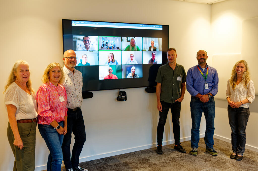 A group of six attendees standing together by a large TV screen displaying participants joining via an online meeting