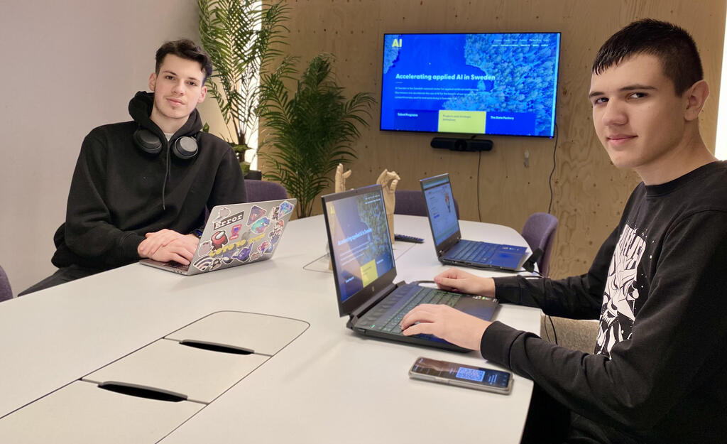 Two young men sitting at a table with their computers, both looking directly into the camera
